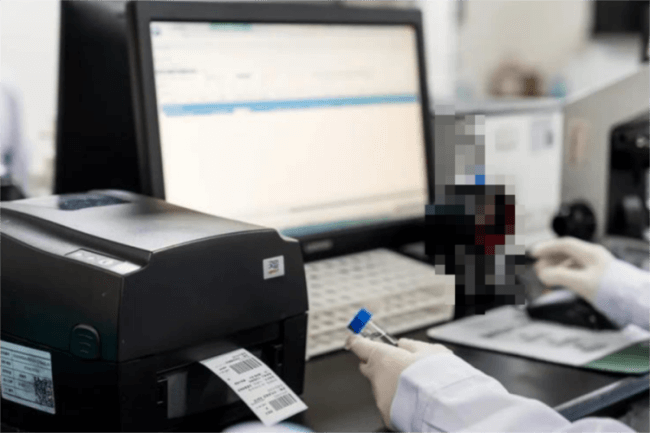 A person operating a εκτυπωτής barcode connected to a computer in a clean workspace.